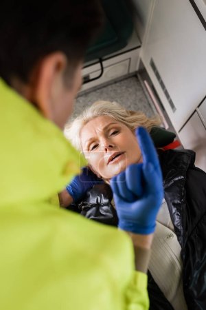 Photo for High angle view of blurred paramedic showing finger while giving first aid to mature woman in emergency vehicle - Royalty Free Image