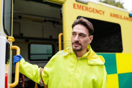 Paramedic in uniform looking away near blurred ambulance car outdoors 