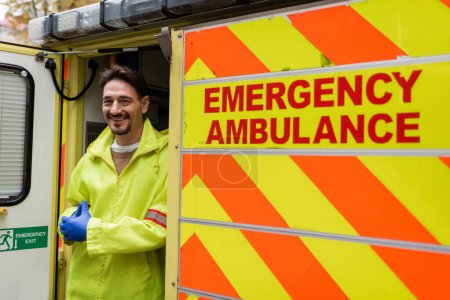 Paramedic in uniform smiling at camera near ambulance vehicle outdoors 