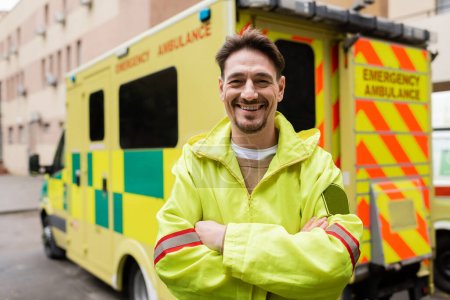 Foto de Sonriente paramédico cruzando brazos y mirando a la cámara cerca de vehículo ambulancia borrosa al aire libre - Imagen libre de derechos
