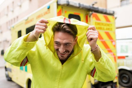 Téléchargez les photos : Un ambulancier souriant coiffé d'une veste près d'une ambulance floue à l'extérieur - en image libre de droit