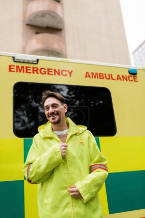 Foto de Sonriendo paramédico usar chaqueta y mirando a la cámara cerca de coche ambulancia al aire libre - Imagen libre de derechos