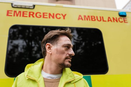 Téléchargez les photos : Vue latérale de l'ambulancier qui regarde loin près de l'ambulance floue auto à l'extérieur - en image libre de droit