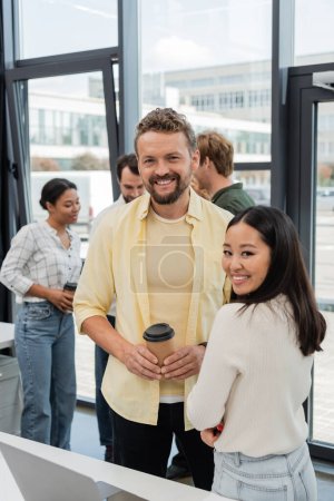 heureux homme d'affaires avec tasse en papier regardant la caméra près de collègue asiatique et l'équipe sur fond
