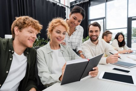 happy businesswoman showing notebook to smiling multiethnic colleagues during meeting in office