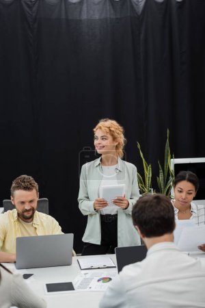 Foto de Mujer de negocios rubia con tableta digital mirando hacia otro lado cerca de colegas interracial trabajando en la oficina - Imagen libre de derechos