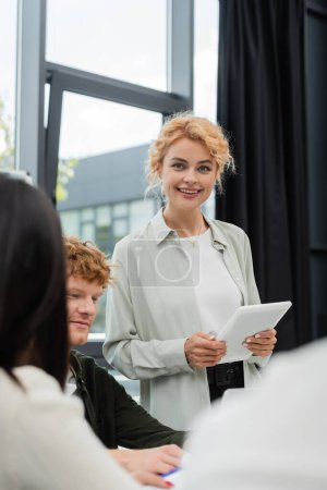 Photo for Blonde businesswoman holding digital tablet and smiling at camera near blurred colleagues - Royalty Free Image