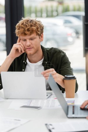 young businessman with red hair thinking near laptop and coffee to go in office