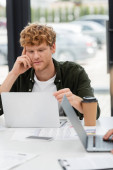 young businessman with red hair thinking near laptop and coffee to go in office t-shirt #623087270