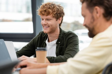 pelirroja hombre de negocios sonriendo cerca de colega trabajando en el ordenador portátil en primer plano borrosa