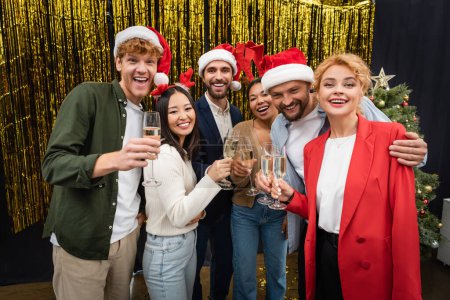 Aufgeregte multiethnische Geschäftsleute mit Weihnachtsmannhüten halten Champagner in der Nähe von Lametta im Büro 