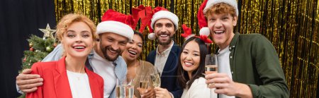 Cheerful interracial business people in santa hats holding champagne near tinsel in office, banner 