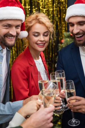Photo for Businesswoman clinking champagne with smiling interracial colleagues in santa hats in office - Royalty Free Image