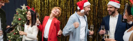 Cheerful interracial business people with champagne dancing and talking during Christmas celebration, banner 