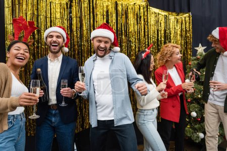 Téléchargez les photos : Des hommes d'affaires multiethniques joyeux dans des chapeaux de Père Noël tenant du champagne près de collègues et tinsel dans le bureau - en image libre de droit