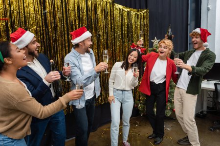 Photo for Positive multicultural business people in santa hats holding champagne near tinsel in office - Royalty Free Image