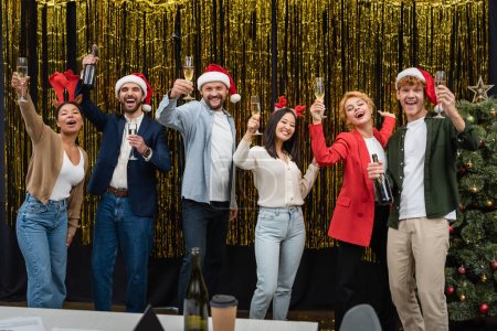Photo for Happy multicultural business people in santa hats holding champagne near Christmas tree and tinsel in office - Royalty Free Image