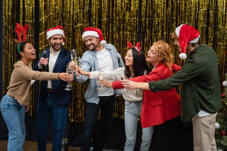 Foto de Gente de negocios multicultural en diademas de Navidad tintineo champán cerca de oropel en la oficina - Imagen libre de derechos
