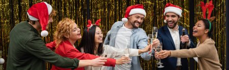 Photo for Positive multiethnic business people in santa hats clinking champagne with colleagues near tinsel, banner - Royalty Free Image