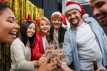 Photo for Positive multicultural business people in santa hats clinking champagne and looking at camera in office - Royalty Free Image