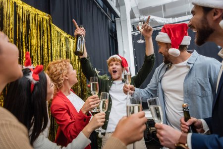 Emocionado hombre de negocios en sombrero de santa celebración de champán mientras se celebra la Navidad con colegas multiétnicos en el cargo 