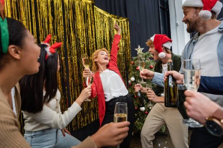 Foto de Empresaria bailando cerca de colegas multiétnicos con champán y árbol de Navidad en la oficina - Imagen libre de derechos