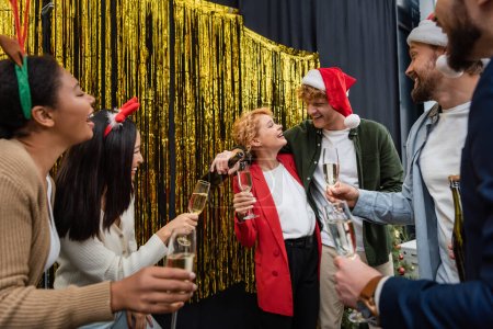Photo for Smiling businessman pouring champagne near interracial colleagues with glasses during Christmas party in office - Royalty Free Image
