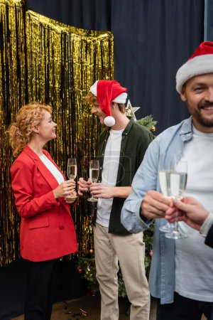 Smiling business people with glasses of champagne talking near tinsel during Christmas party in office 