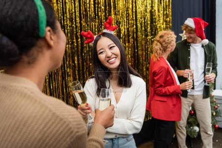 Smiling asian businesswoman holding champagne and talking to multiracial colleague during Christmas party in office 