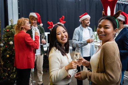 Foto de Mujeres de negocios multiétnicas sonrientes en diademas de Navidad sosteniendo champán y mirando a la cámara en la oficina - Imagen libre de derechos
