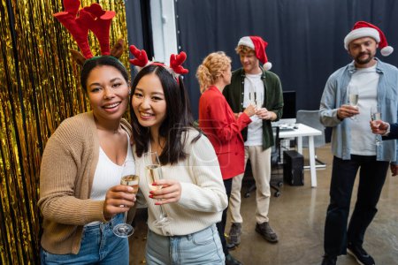 Téléchargez les photos : Femmes d'affaires interraciales positives dans les bandeaux de Noël tenant du champagne près de tinsel au bureau - en image libre de droit