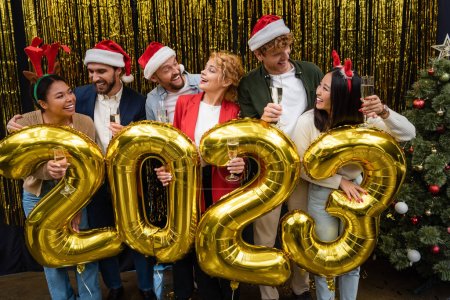 Positive multikulturelle Geschäftsleute in Weihnachtsmützen mit Champagner in der Nähe von Luftballons in Form von 2023 im Amt 