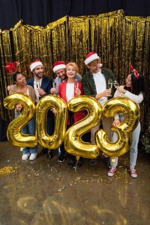 Foto de Gente de negocios interracial sosteniendo copas de champán cerca de globos en forma de 2023 y oropel en la oficina - Imagen libre de derechos