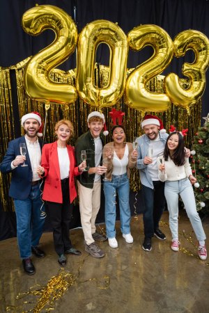 Positive multikulturelle Geschäftsleute mit Weihnachtsmützen und Champagner in der Nähe von Luftballons im Jahr 2023 im Amt 
