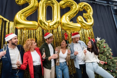 Lächelnde multiethnische Geschäftsleute mit Champagner und Luftballons in Form von 2023-Schildern im Amt 