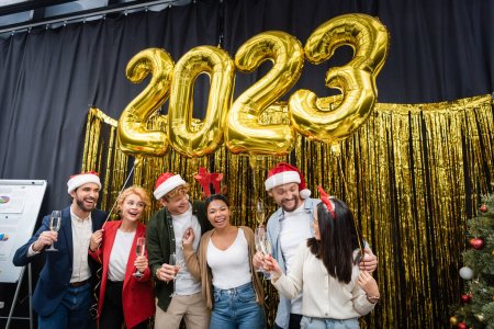 Des hommes d'affaires multiethniques regardant un collègue asiatique avec du champagne près de ballons en forme de 2023 signes au bureau 