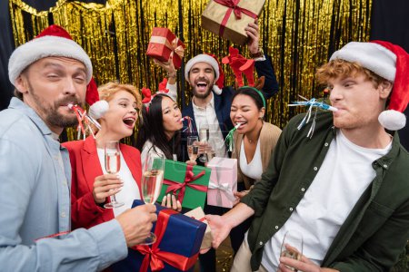Smiling multiethnic business people in santa hats holding champagne and gifts near tinsel during party in office 