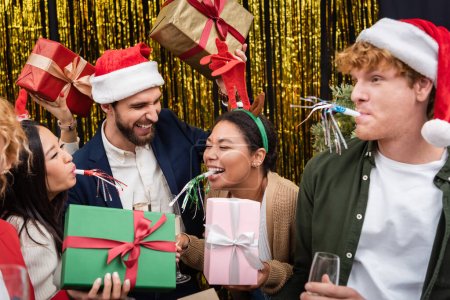 Multiracial businesswoman in Christmas headband holding present and blowing party horn near colleagues in office 
