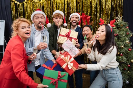 Photo for Multiethnic business people with gifts and champagne blowing party horns during Christmas corporate party in office - Royalty Free Image