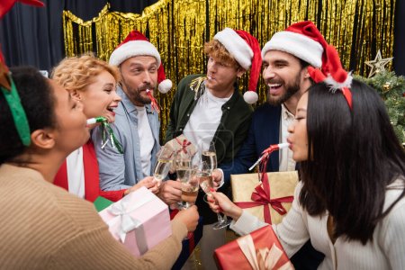 Multiethnische Geschäftsleute mit Weihnachtsmannhüten blasen bei der Weihnachtsfeier im Büro Parteihupen und klirren mit Kollegen Champagner 