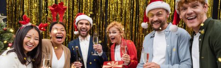 Téléchargez les photos : Des hommes d'affaires interraciaux positifs avec des verres à champagne debout sous des confettis pendant la fête de Noël au bureau, bannière - en image libre de droit