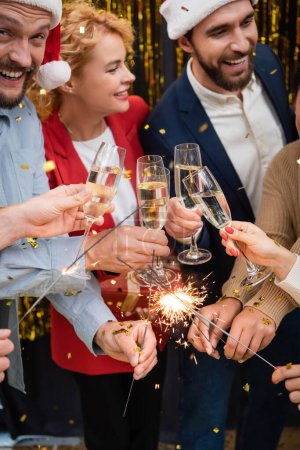 Des hommes d'affaires multiethniques joyeux tenant des verres de champagne et des étincelles pendant la fête de Noël au bureau 