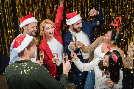 Téléchargez les photos : Des hommes d'affaires interraciaux positifs tenant du champagne et dansant sous des confettis pendant la fête de Noël au bureau - en image libre de droit