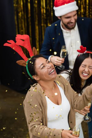 High angle view of multiracial businesswoman holding champagne under confetti during Christmas party in office 