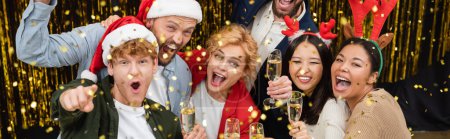 Foto de Alegre gente de negocios multiétnicos en sombreros de santa celebración de champán bajo confeti durante la fiesta corporativa de Navidad en la oficina, pancarta - Imagen libre de derechos