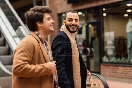 cheerful bearded man with shopping bags looking at gay partner on city street