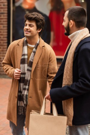 Foto de Feliz gay hombre en beige abrigo mirando lejos cerca barbudo novio con compras bolsas - Imagen libre de derechos
