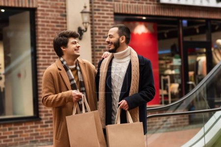 Foto de Elegante pareja gay con bolsas de compras sonriendo el uno al otro cerca de la tienda en un fondo borroso - Imagen libre de derechos