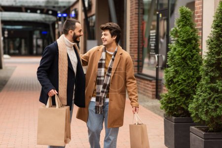 excited gay men with shopping bags looking at each other near green plants on street