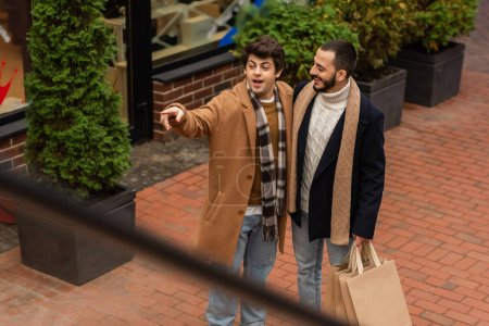 Foto de Alto ángulo vista de asombrado gay hombre mirando lejos y apuntando con dedo cerca sonriente novio con bolsas de compras - Imagen libre de derechos